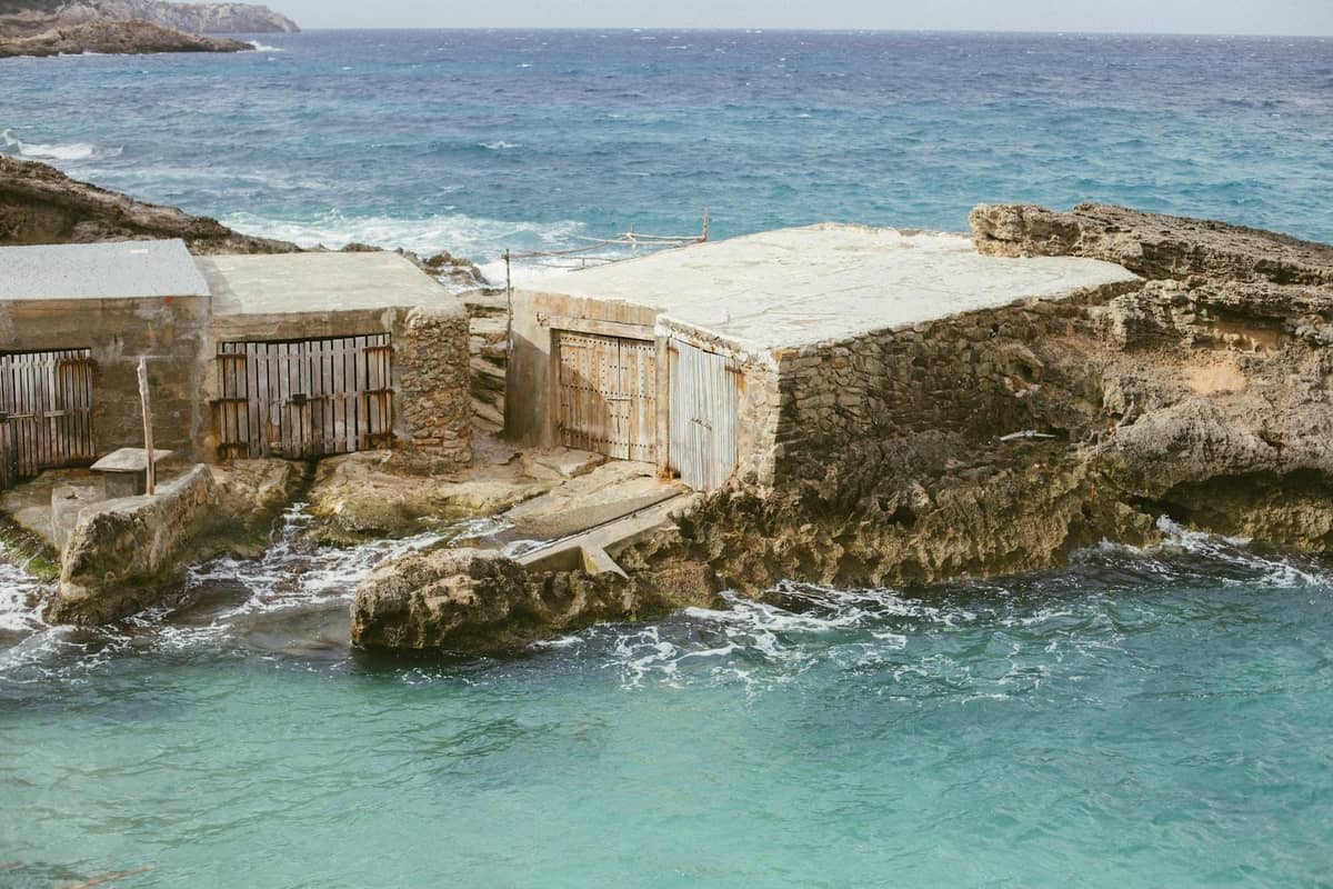 Traditionelle hölzerne Fischerhütten am felsigen Ufer am klaren türkisfarbenen Meer in Mallorca, mit Wellen, die gegen die Eingänge spritzen.