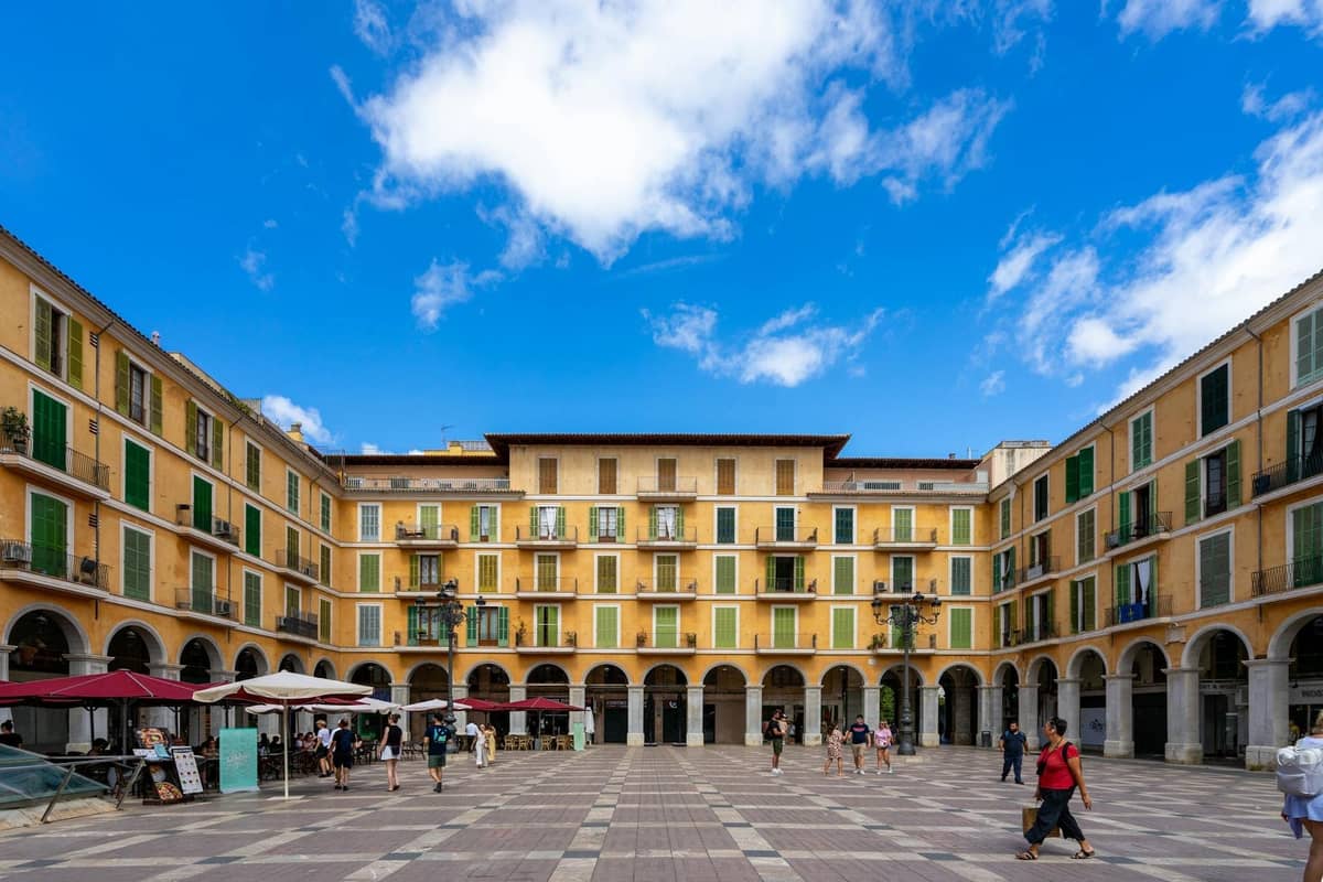 Maluwag na plaza sa Palma de Mallorca na may mga dilaw na gusali, berdeng mga shutter, arched na daanan, mga street cafe at mga taong naglalakad sa ilalim ng asul na langit.