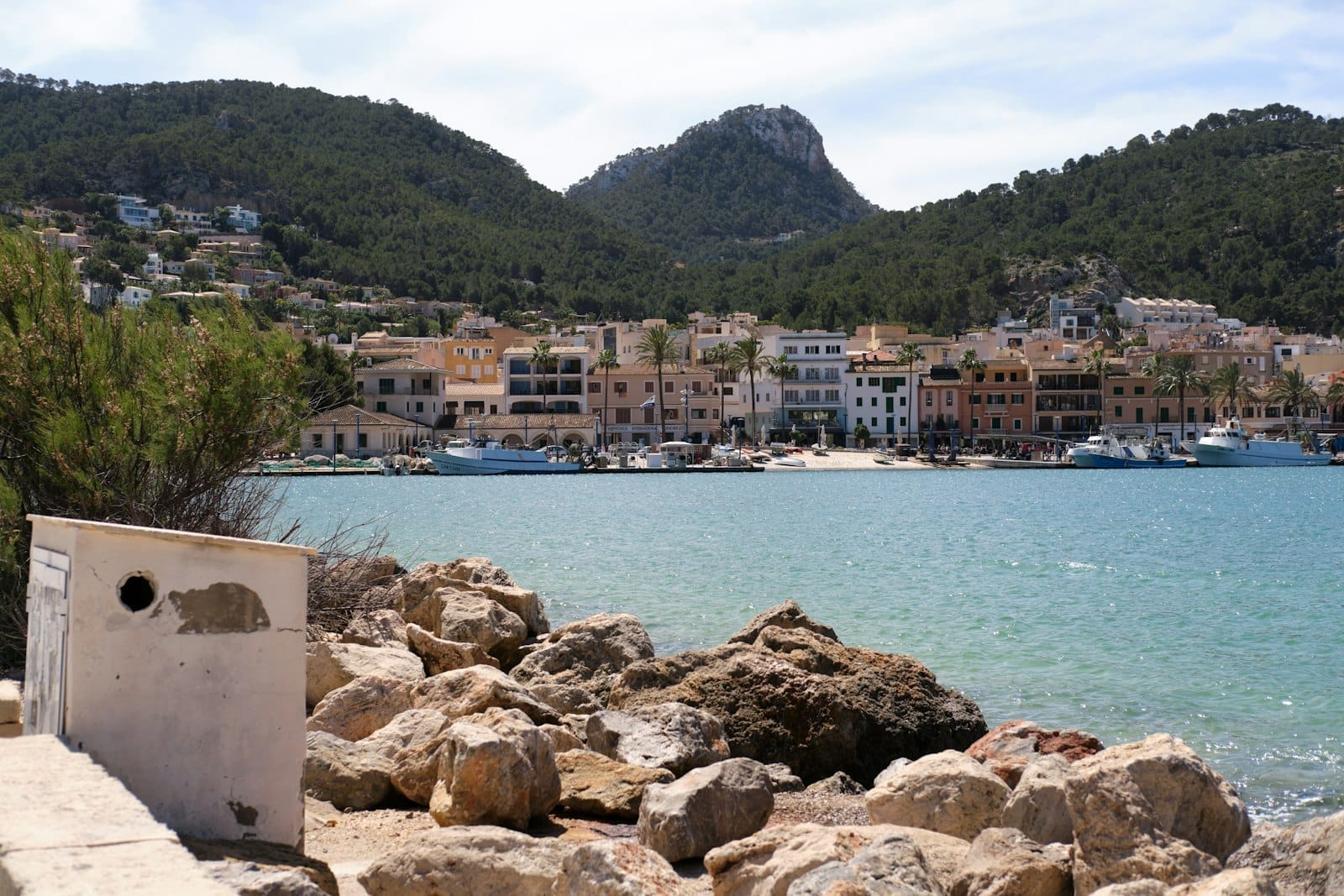 Vista do cais de Port Andratx em Mallorca com edifícios coloridos, barcos atracados, costa rochosa e colinas verdes sob um céu luminoso.