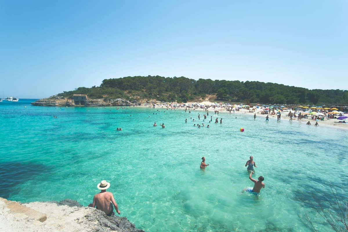 Pantai Mallorca yang ramai di musim panas dengan air berwarna biru kehijauan yang jernih, banyak orang berenang, berjemur, dan bersantai di bawah langit biru yang cerah.