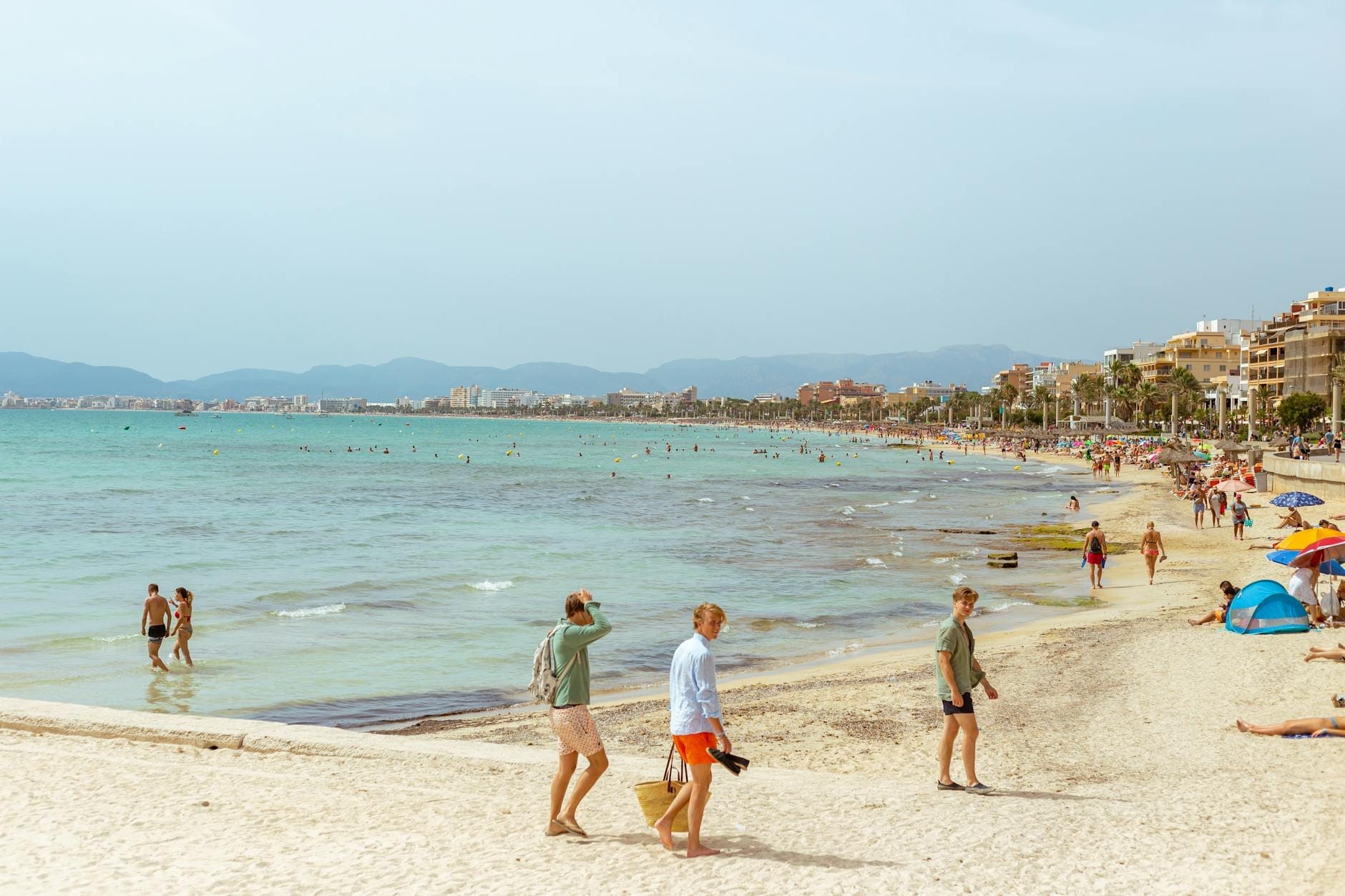 Pessoas caminhando e relaxando em uma praia de areia de Maiorca com água turquesa, guarda-sóis e a agitação típica do verão em julho ou agosto.