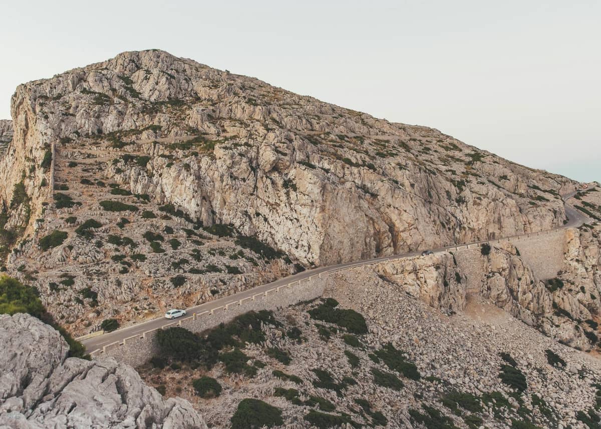Jalan berkelok-kelok dengan sebuah mobil yang melintasi lanskap pegunungan berbatu di Mallorca, menggambarkan perjalanan dengan mobil di pedalaman pulau yang terjal.