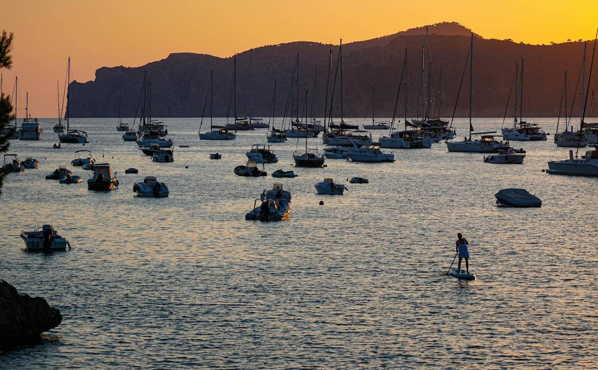Barcos y yates anclados al atardecer en las tranquilas aguas del Mediterráneo con colinas al fondo, evocando el ambiente de las fiestas en barco al anochecer en Mallorca.