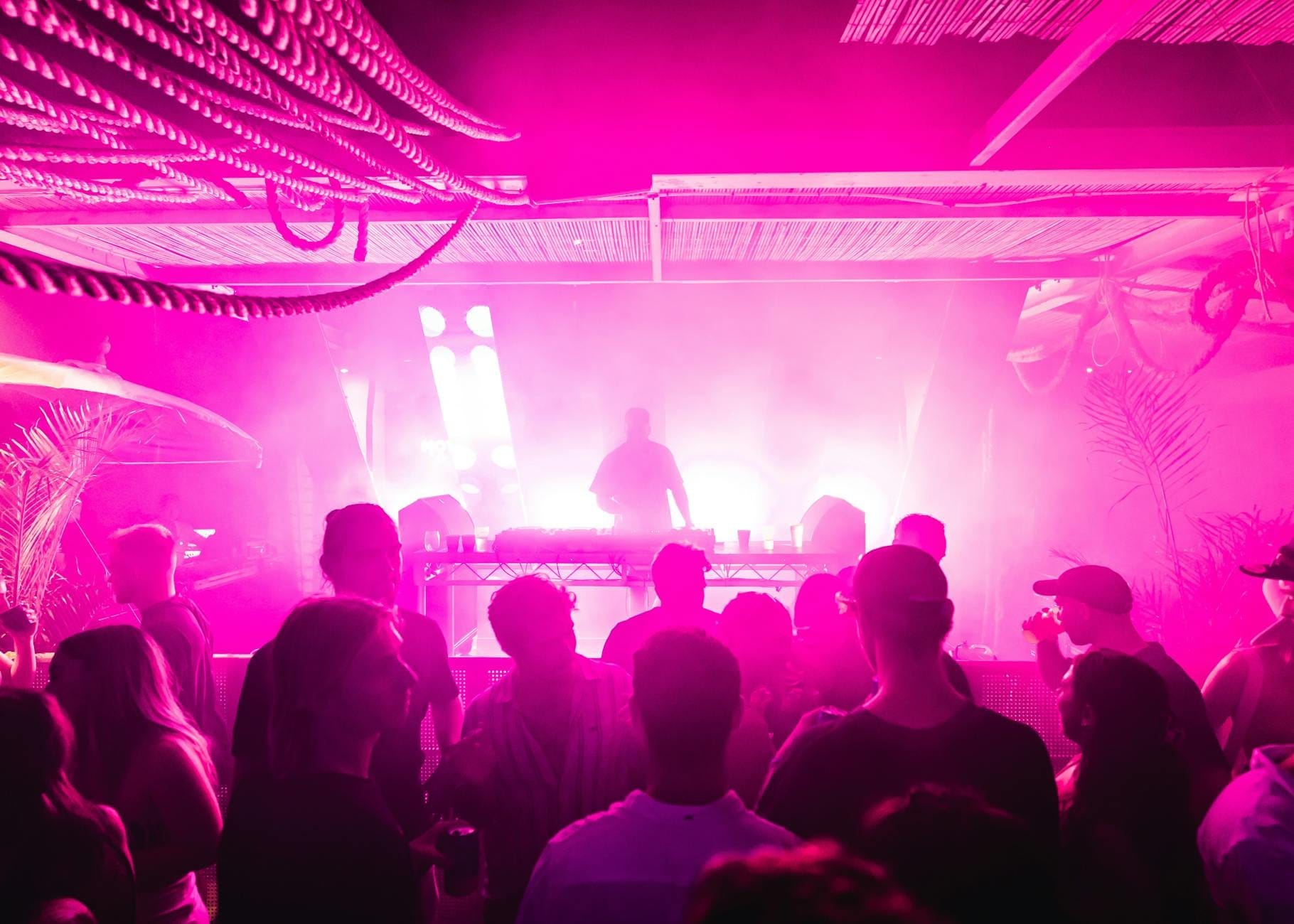 Multidão curtindo uma balada animada ou festa de praia em Maiorca com iluminação rosa e roxa e um DJ se apresentando no palco.