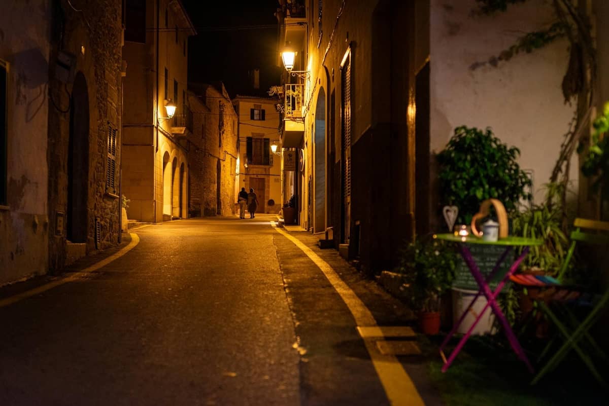 Una calle estrecha y cálidamente iluminada de noche con edificios históricos de piedra, farolas y una acogedora mesa de café con sillas junto a la pared.