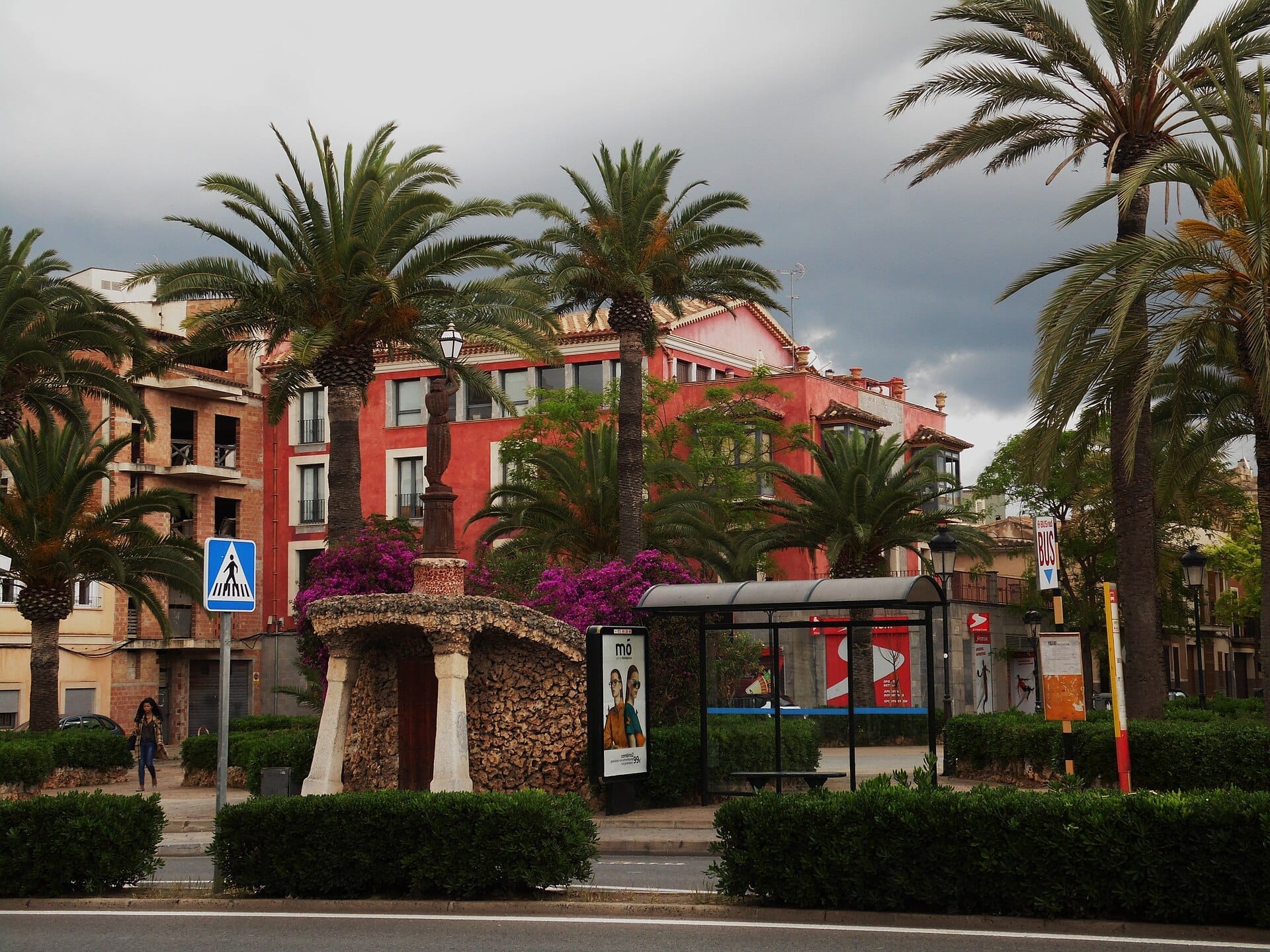 Vista di una strada a Manacor con palme, un monumento in pietra, edifici rosa e una fermata dell'autobus coperta sotto un cielo nuvoloso.