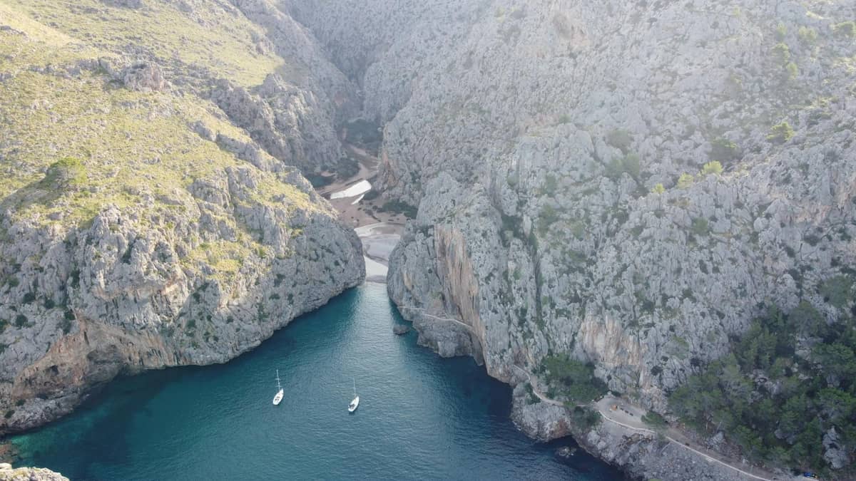 Una drammatica gola rocciosa con acque turchesi, scogliere ripide e due piccole imbarcazioni ancorate in basso, che mostrano i paesaggi naturali incontaminati di Maiorca.