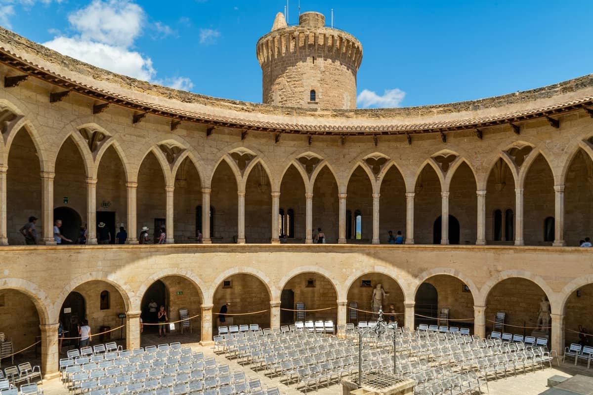 Cortile in pietra di un castello circolare medievale con portici ad arco e una torre centrale sotto un cielo azzurro a Mallorca.