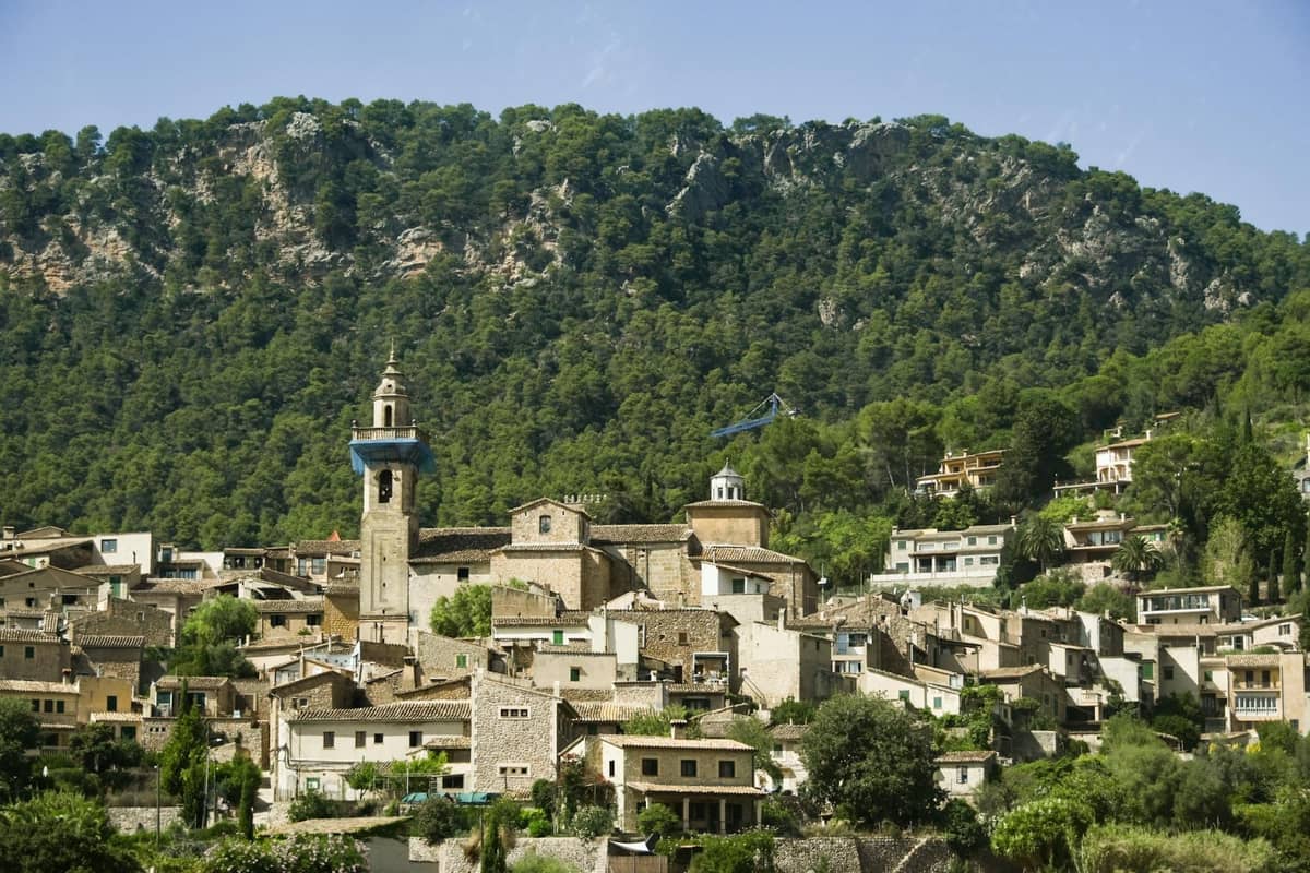 Villaggio in pietra con campanile adagiato sullo sfondo lussureggiante della sierra Tramuntana a Mallorca sotto un cielo azzurro.