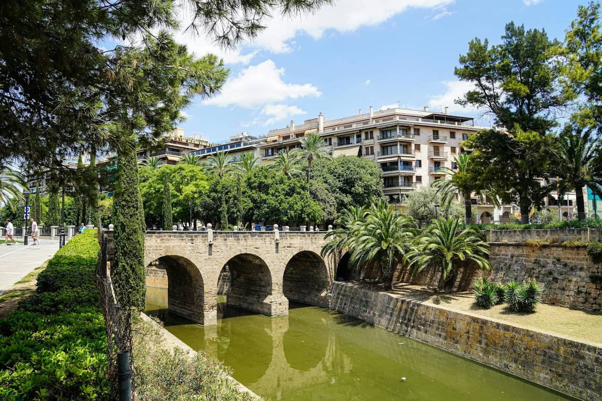 Ponte ad arco in pietra su un canale, con rigogliosa vegetazione ed edifici sullo sfondo in un paese di Mallorca.