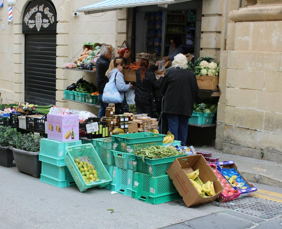 Pequeno grupo de pessoas comprando produtos frescos em um mercado de rua ao ar livre em uma vila mediterrânea de pedra.