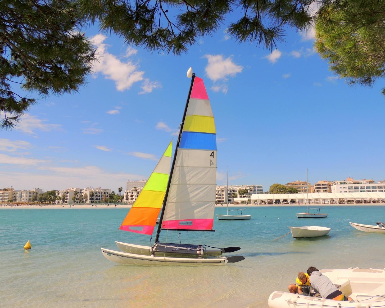 Veleiro colorido em uma praia calma em Mallorca, água turquesa cristalina, crianças brincando à beira-mar e céu azul com nuvens esparsas.