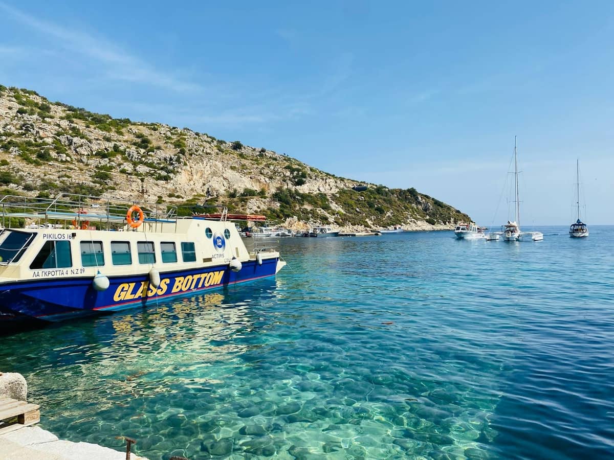Isang bangkang may salaming ilalim na nakaangkla malapit sa dalampasigan na may malinaw na turquoise na tubig, mga batuhang burol, at mga layag sa likod sa Mallorca.