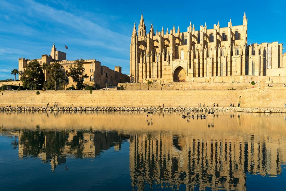 Malawak na tingin ng medyebal na katedral ng Mallorca, mga pader ng lungsod, at salamin ng tubig sa maliwanag na sikat ng araw, na nagpapakita ng kahanga-hangang makasaysayang arkitektura na angkop para sa paglalakbay ng pamilya.