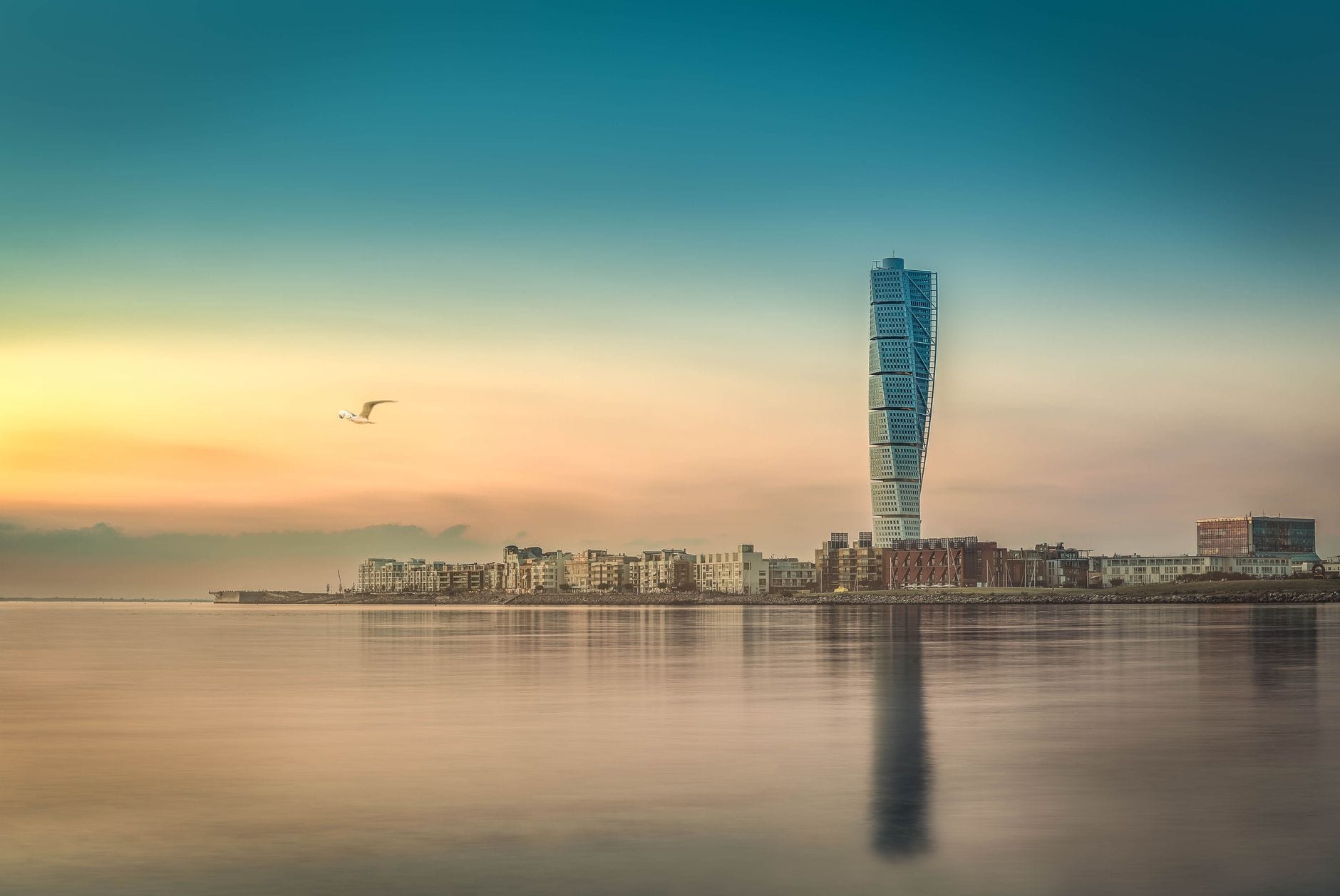 Vista al tramonto del grattacielo Turning Torso di Malmö e degli edifici sul lungomare, riflessi nell'acqua calma con un gabbiano in volo.