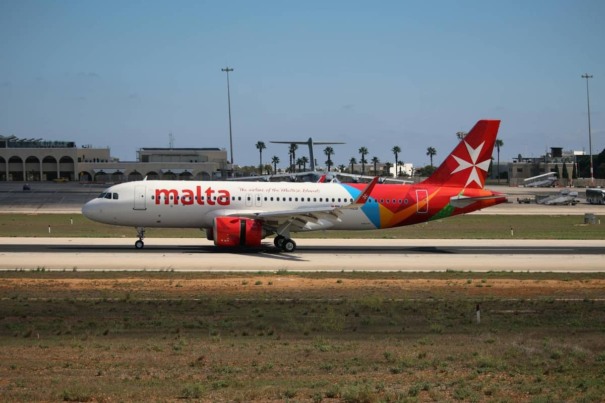 Eroplano ng Air Malta sa runway ng Malta International Airport na may terminal at mga puno ng palma sa likod