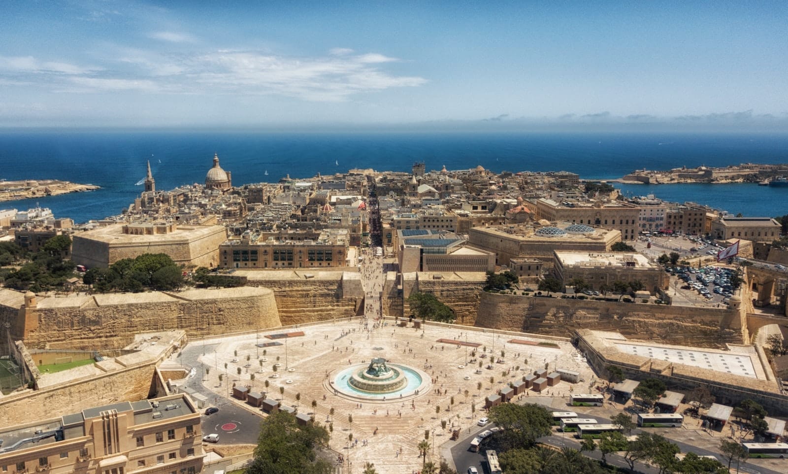 Vista aérea panorâmica de Valletta, Malta, com edifícios históricos de calcário, uma grande rotatória com fonte e o Mar Mediterrâneo ao fundo sob céu azul.