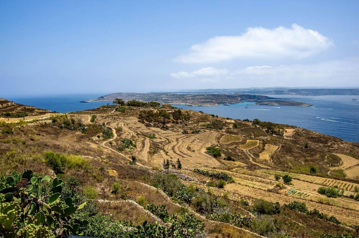 Malawak na tanawin ng mga terraced na burol ng Gozo na may Mediterranean Sea at Comino Island sa malayo sa ilalim ng asul na langit.