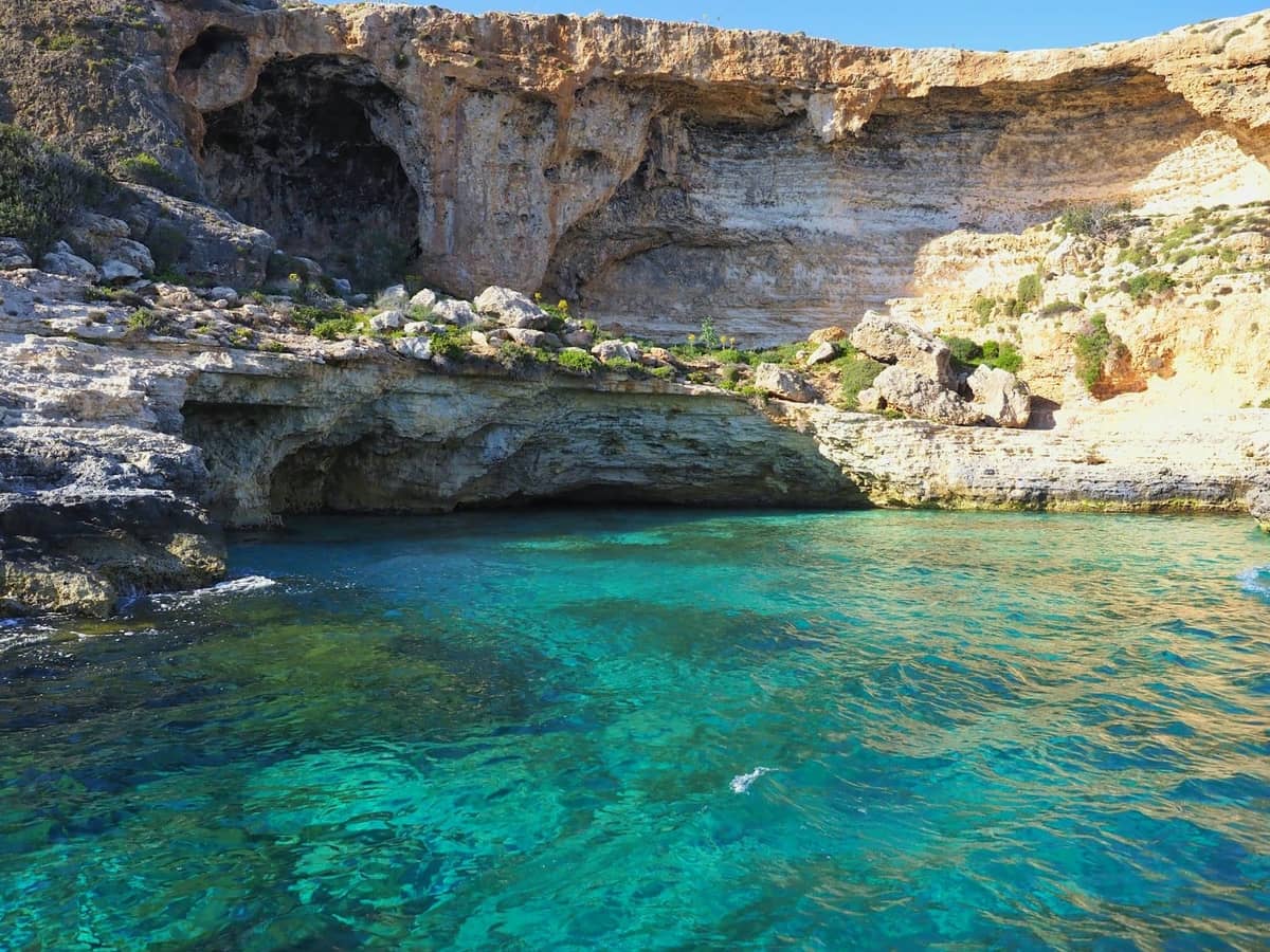 Чистые голубые воды лагуны со скалистыми утёсами и видимыми морскими пещерами на острове Комино, Мальта, под солнечным небом.