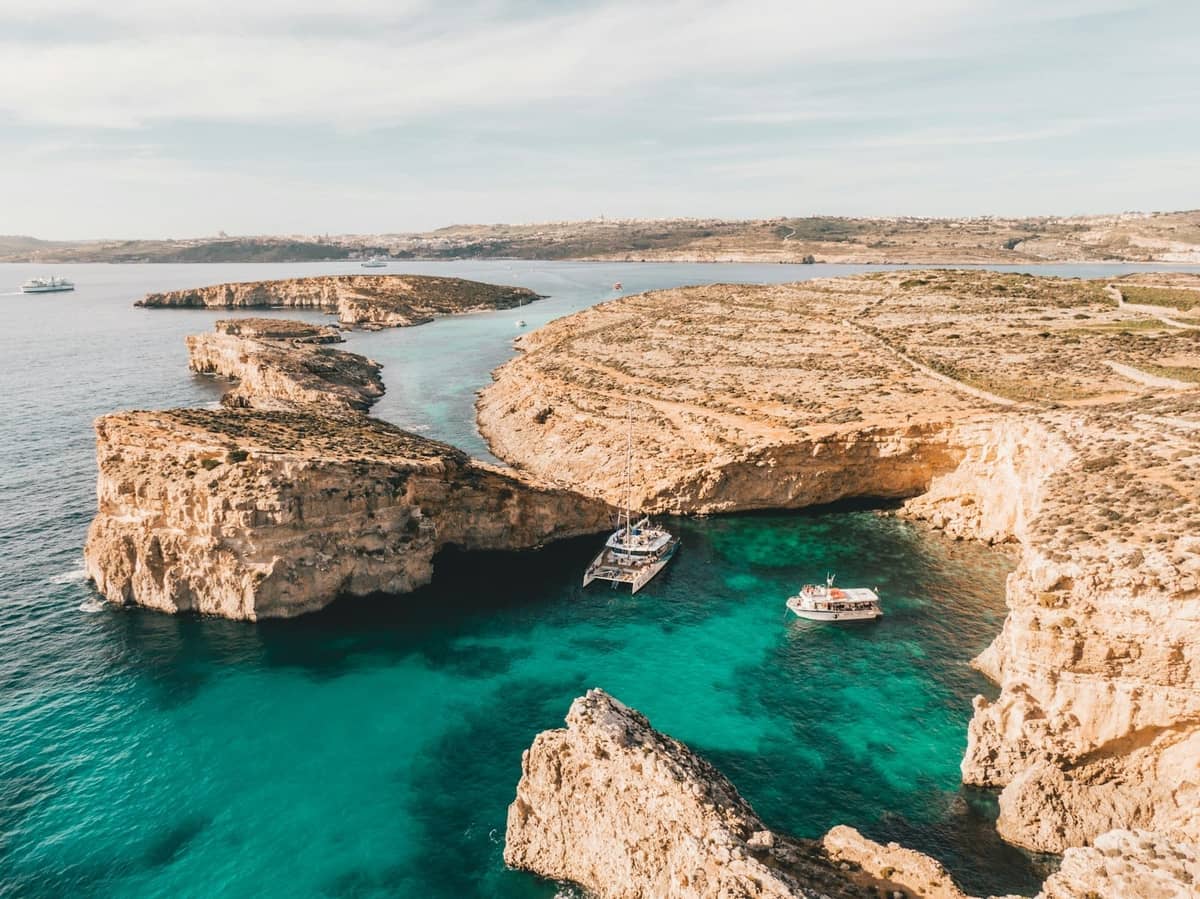 Aerial view of Malta’s rugged coastline with turquoise clear water and boats anchored in a picturesque cove, showcasing top diving conditions.
