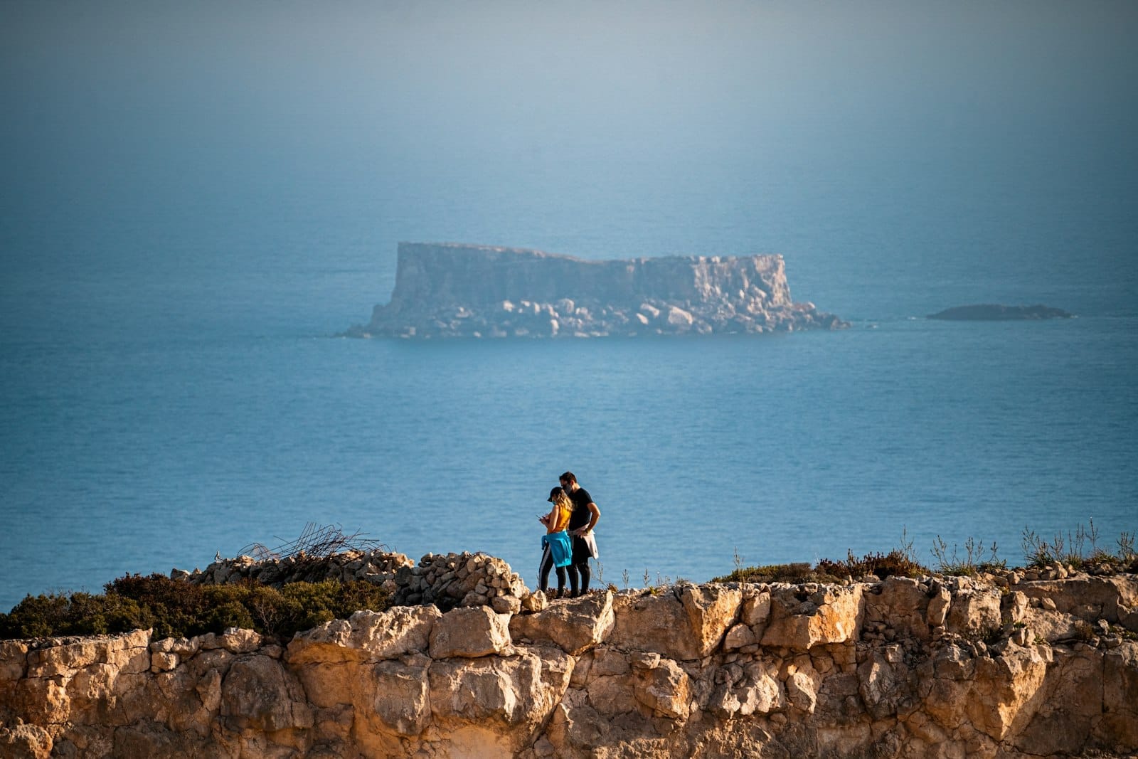 Casal bem próximo em uma falésia, contemplando o mar com uma ilha rochosa ao fundo sob céu limpo em Malta.