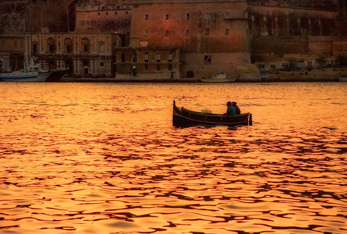 Piccola barca con una coppia a bordo al tramonto, con acqua dorata e gli storici edifici in pietra di Malta sullo sfondo.
