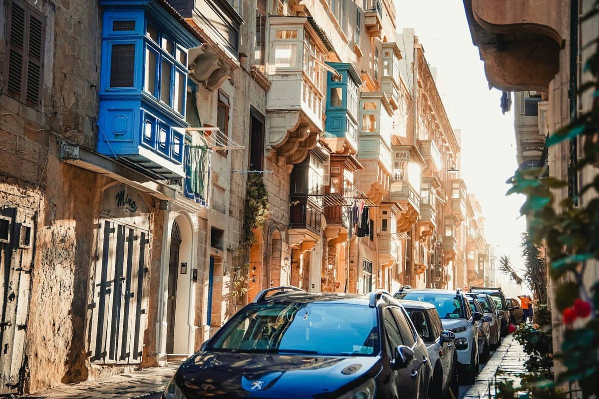 Strada soleggiata a La Valletta fiancheggiata da edifici barocchi color miele e tradizionali balconi maltesi, con auto parcheggiate lungo la strada stretta.