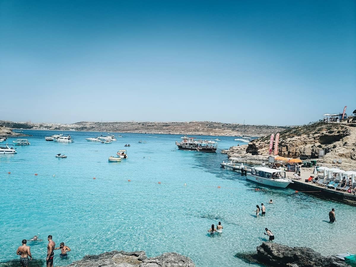 Krystalicznie turkusowe wody Blue Lagoon z pływakami, skalistym brzegiem i zakotwiczonymi łodziami na Comino, Malta, pod jasnym błękitnym niebem.