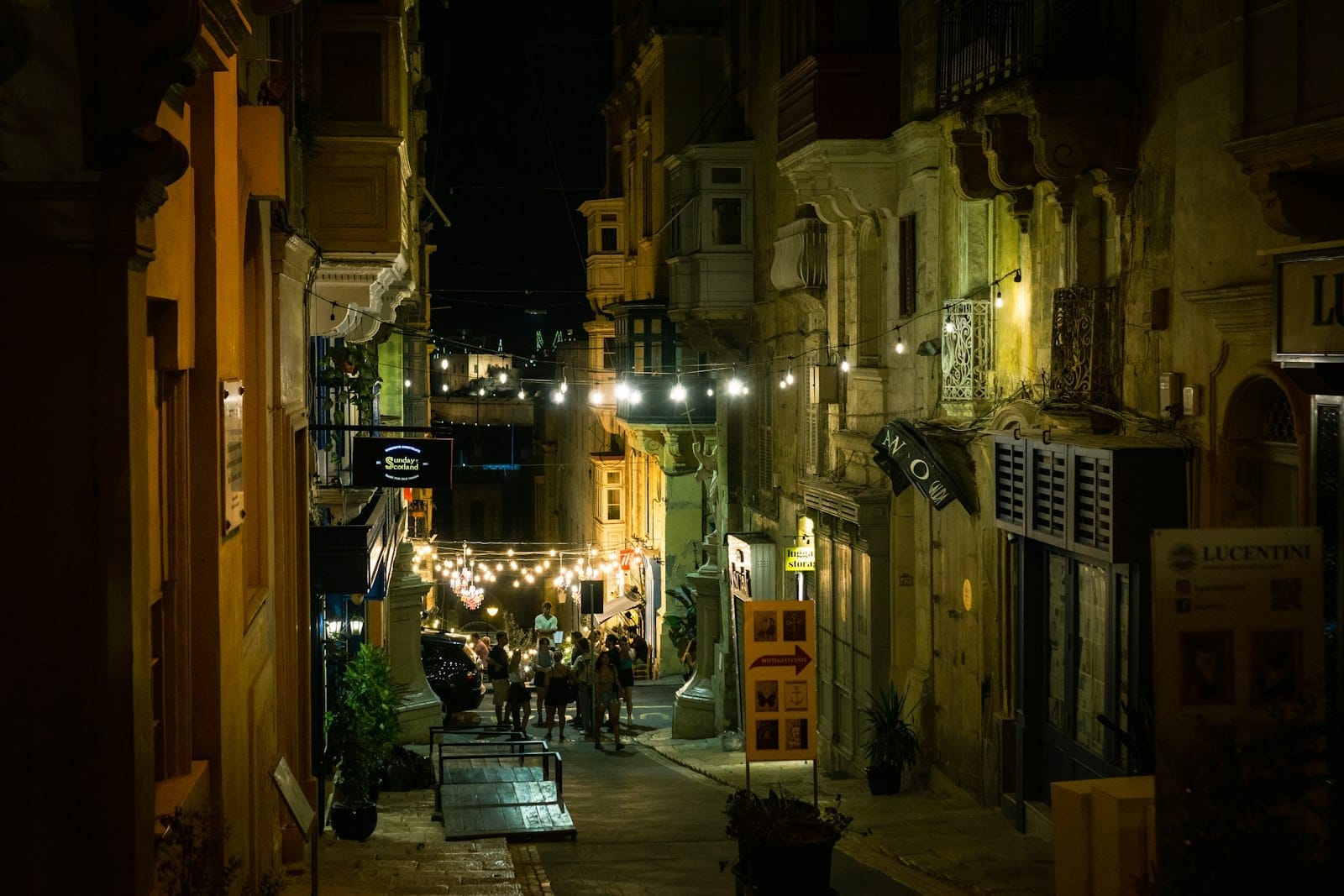 Rua estreita em Valletta à noite com cordões de luzes acesas, grupos de pessoas reunidas em frente a bares e prédios históricos dos dois lados.