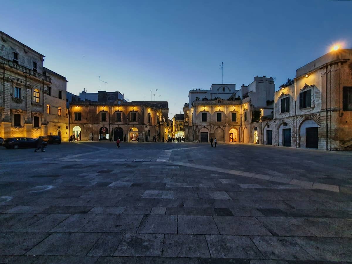 Một quảng trường đá rộng lớn ở Valletta lúc hoàng hôn, được bao quanh bởi những tòa nhà Baroque được chiếu sáng nhẹ nhàng bằng ánh đèn buổi tối ấm áp.