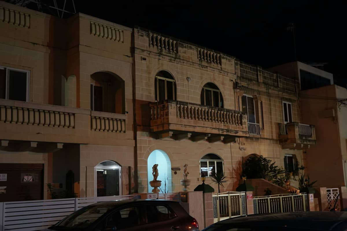Night view of a modest guesthouse or budget hotel building with balconies and cars parked outside, typical of Malta accommodation.