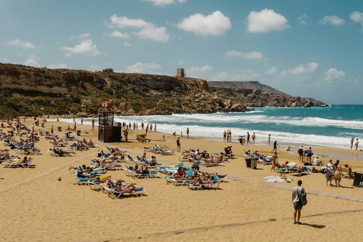 Popular sandy Malta beach on a sunny day, with many people sunbathing and swimming, and a rocky headland in the background.