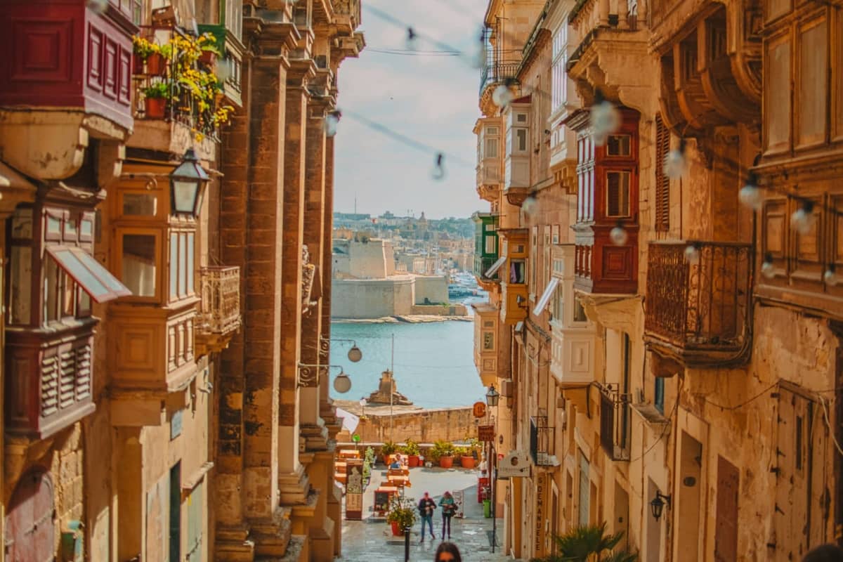 Sunlit street in Valletta, Malta with historic Baroque buildings, colorful balconies, and a distant view of the harbor and sea.
