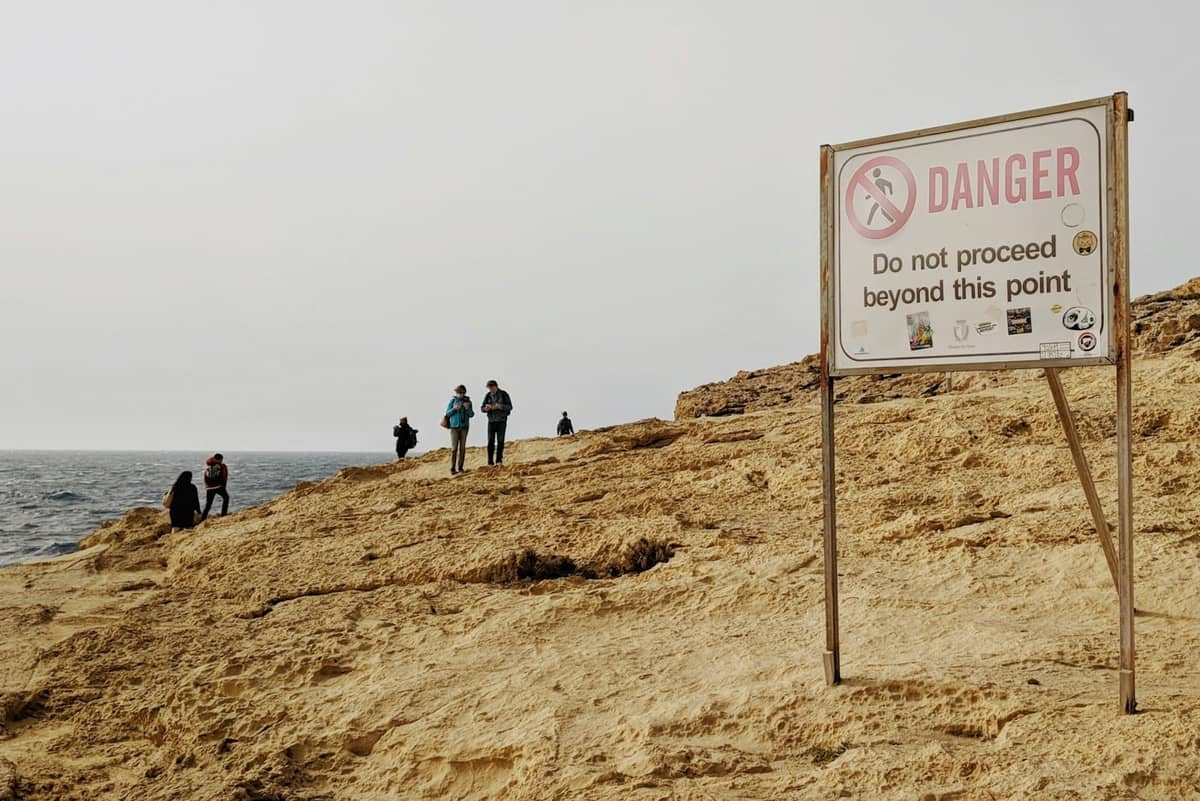 Felsige Küste Maltas mit einem großen 'Danger'-Warnschild nahe der Klippe, mehrere Personen stehen sicher vom Rand entfernt.
