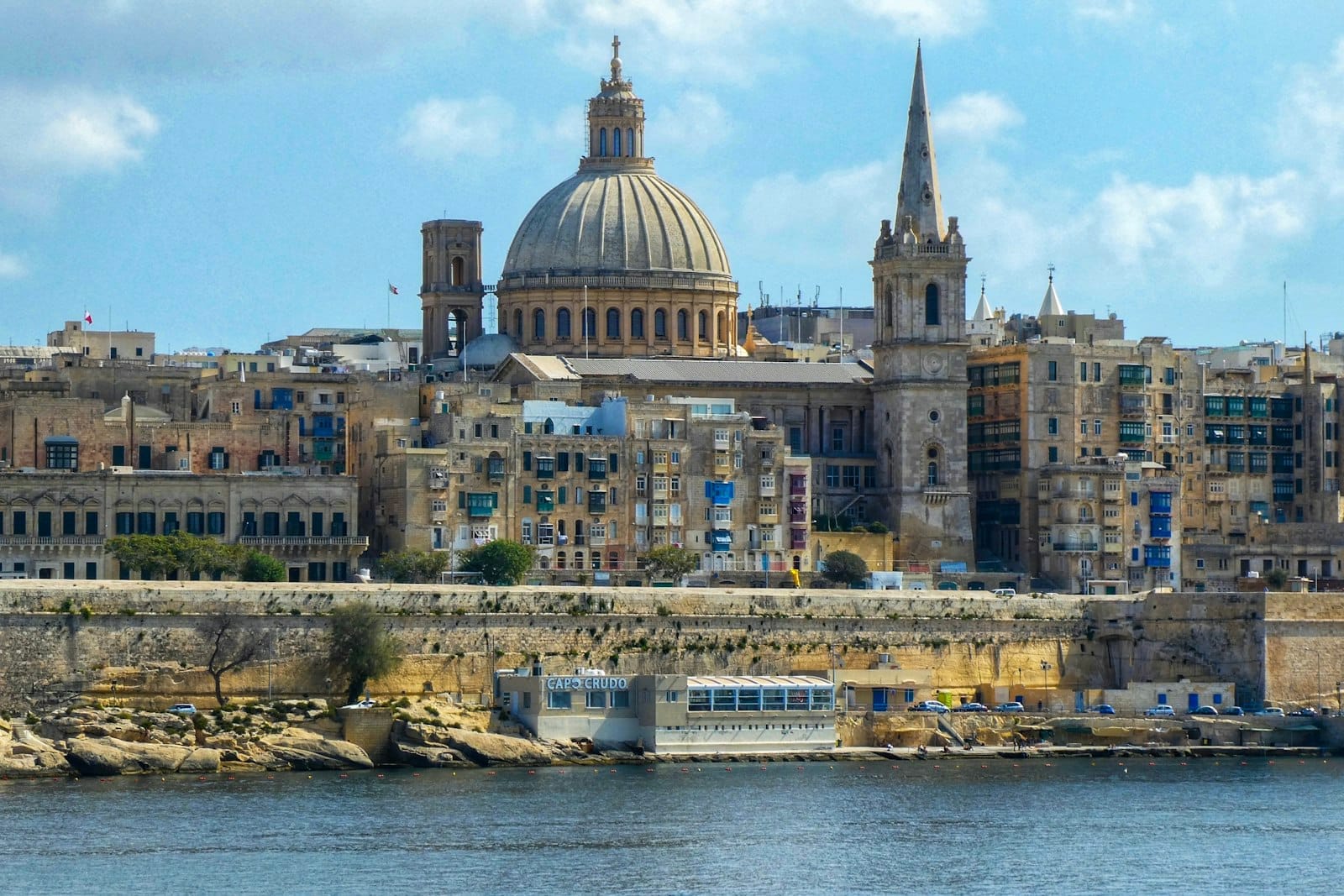 A stunning panoramic view of Valletta, Malta’s capital, with its iconic dome of St. Paul's Cathedral and spire, historic limestone buildings, and a bright blue sky above the waterfront.