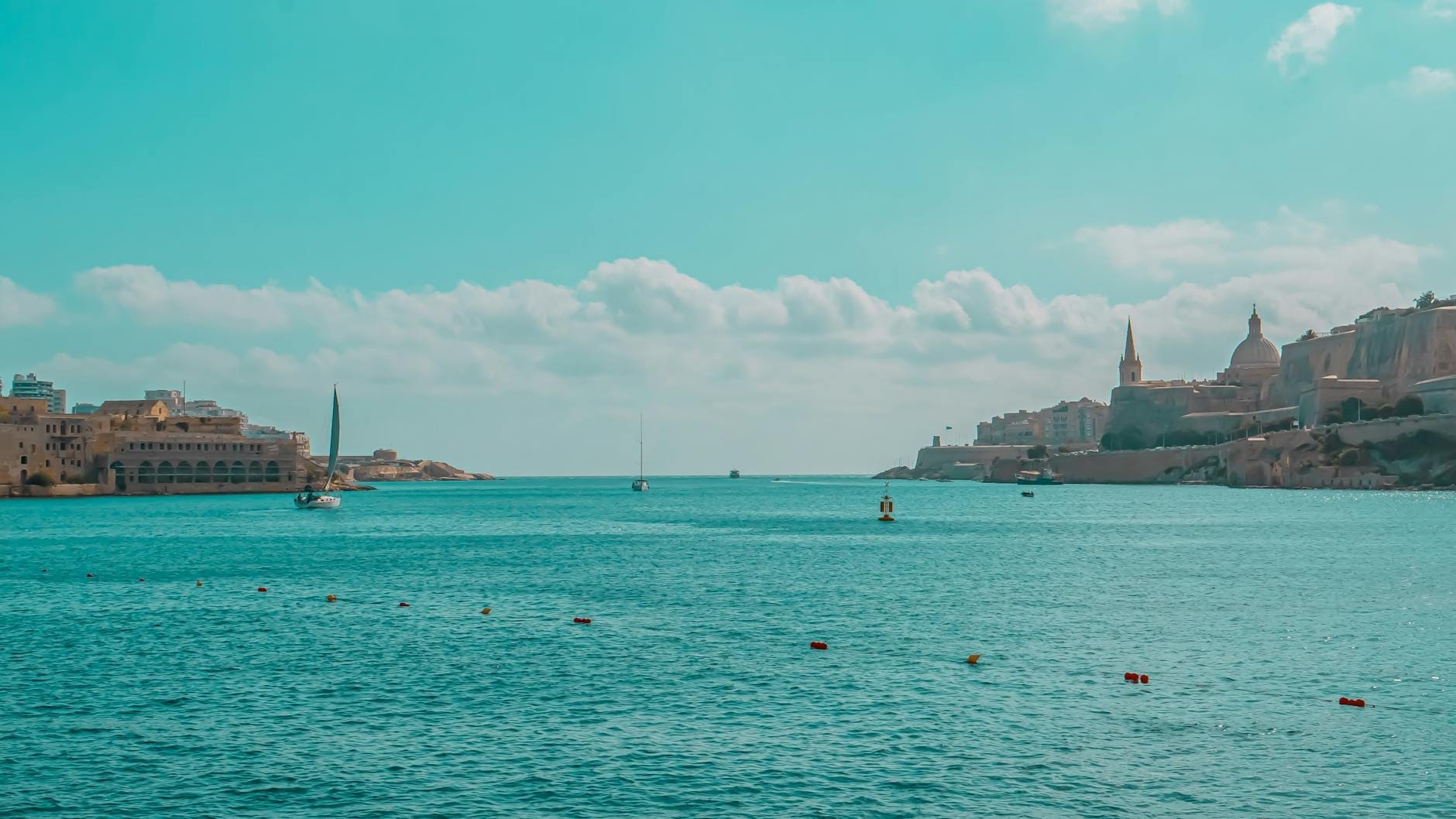 Mar turquesa calmo com veleiros e o histórico skyline de Valletta sob um céu azul, capturando o ponto de partida de Malta para a ferry à Sicília.