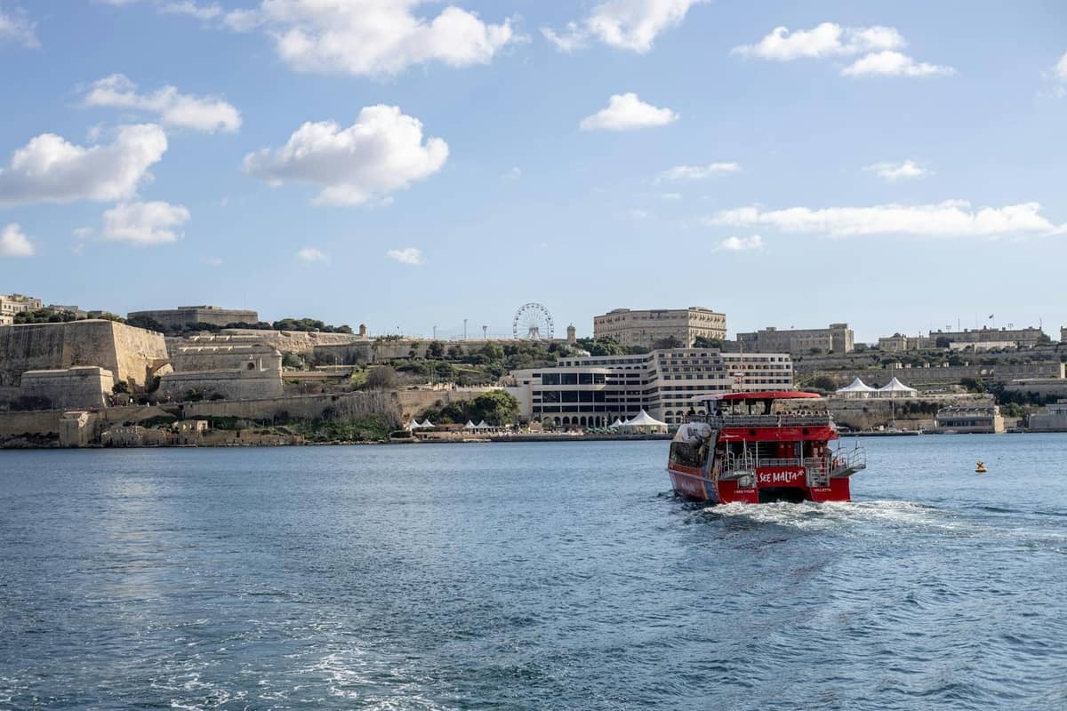 Un ferry rojo cruzando el puerto de Malta con edificios históricos de piedra y una noria al fondo en un día soleado.