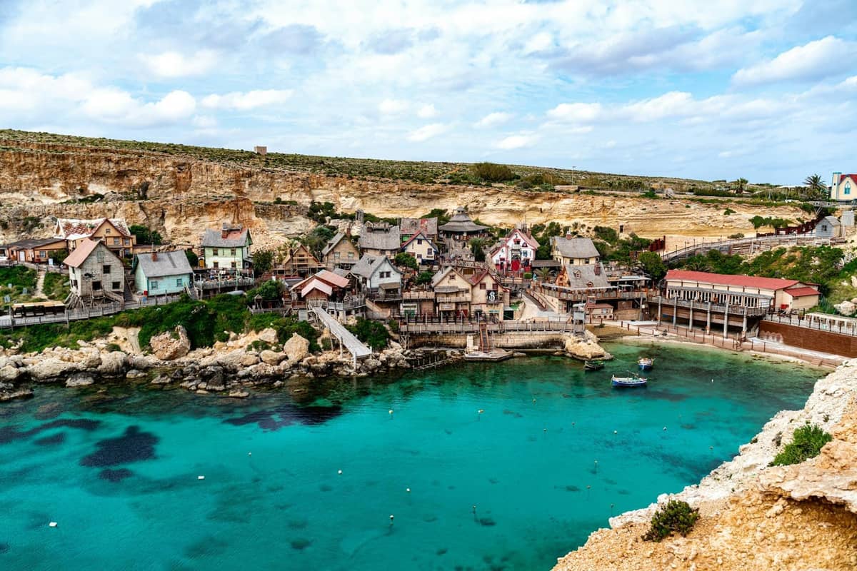 Szeroki widok na Popeye Village z kolorowymi drewnianymi budynkami nad turkusową Anchor Bay na Malcie pod błękitnym niebem.
