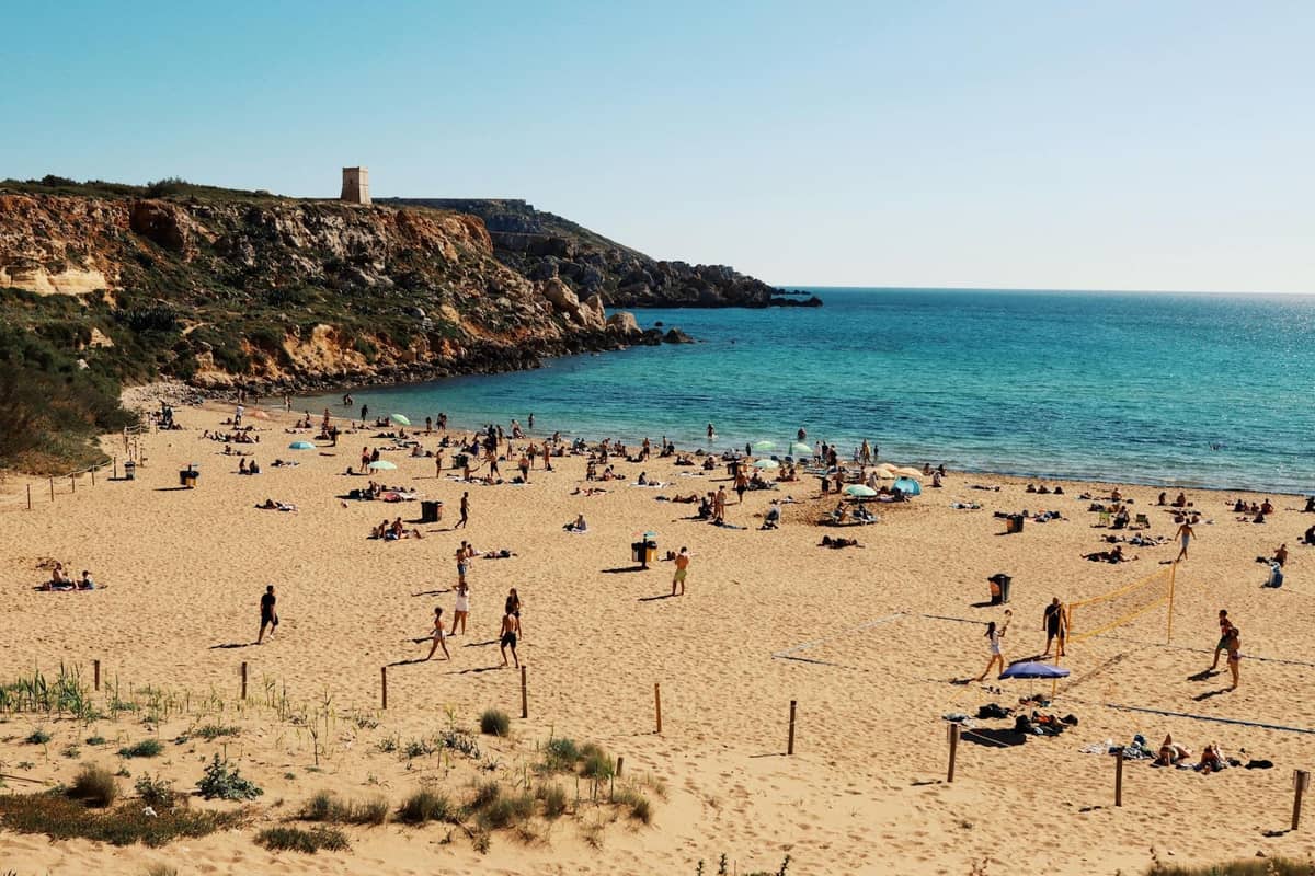 Szeroka piaszczysta plaża na Malcie z rodzinami i bawiącymi się dziećmi, plażowiczami opalającymi się oraz płytkimi, błękitnymi wodami. W tle widoczne są skaliste klify i wieża strażnicza.