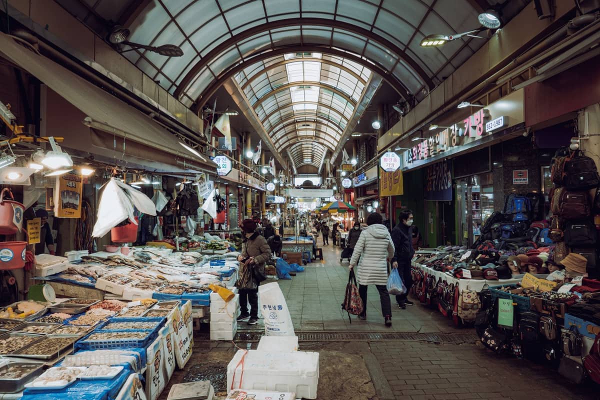 Corredor coberto dentro do Mercado Mangwon em Seul com bancas de frutos do mar frescos, mochilas à venda e compradores carregando sacolas sob as luzes brilhantes do mercado.
