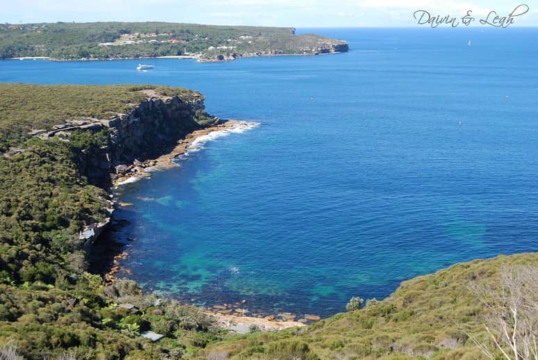 Vista del Manly Scenic Walkway sobre un cabo de arenisca cubierto de matorral, una cala rocosa clara y el océano azul intenso hacia una costa acantilada lejana.