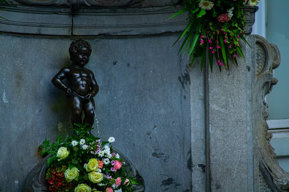 Estátua do Manneken Pis em Bruxelas, uma pequena fonte de bronze em forma de menino sobre pedra com flores coloridas decorando a base.