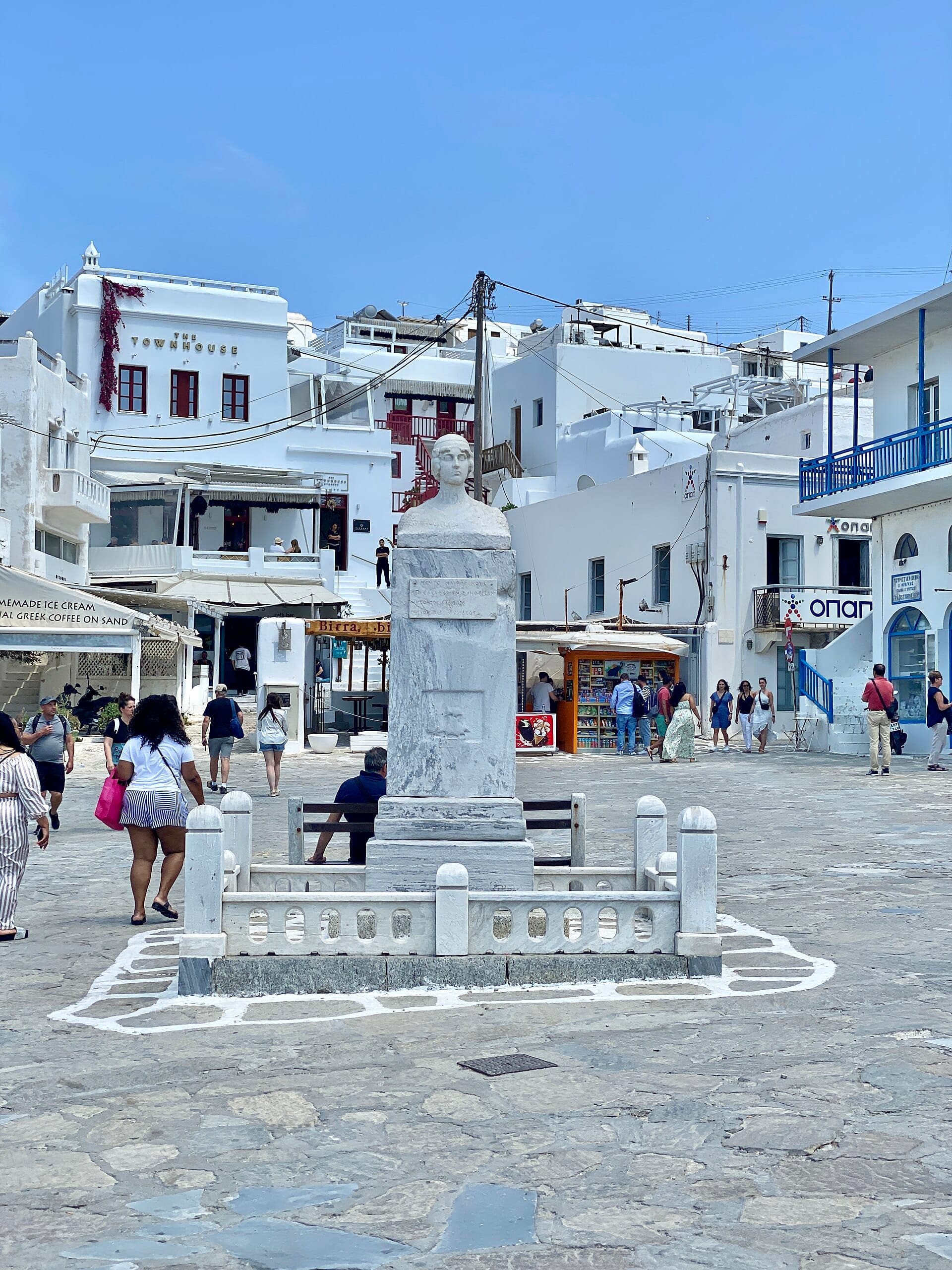 Estatua de mármol de Manto Mavrogenous en el centro de la plaza, rodeada de edificios cicládicos blancos y personas explorando Mykonos Town.