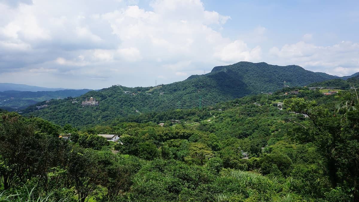 Üppig grüne Hügel von Maokong mit vereinzelten Gebäuden und Tempeln, Panoramablick unter blauem Himmel mit Wolken – das Teeanbaugebiet in Taipeis Bergen.