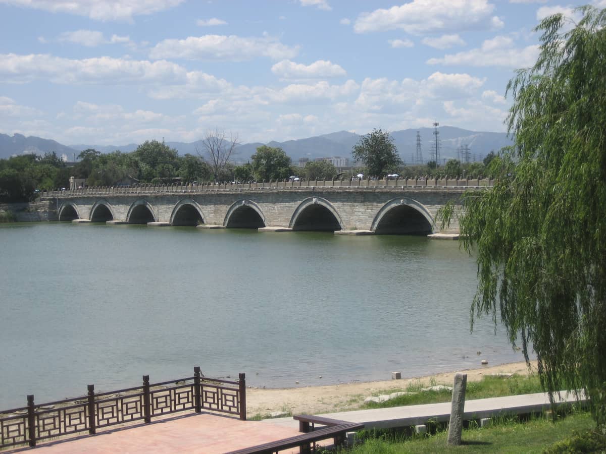 Die Marco-Polo-Brücke wölbt sich über einen ruhigen Fluss mit üppigem Grün und fernen Bergen unter einem teilweise bewölkten blauen Himmel.