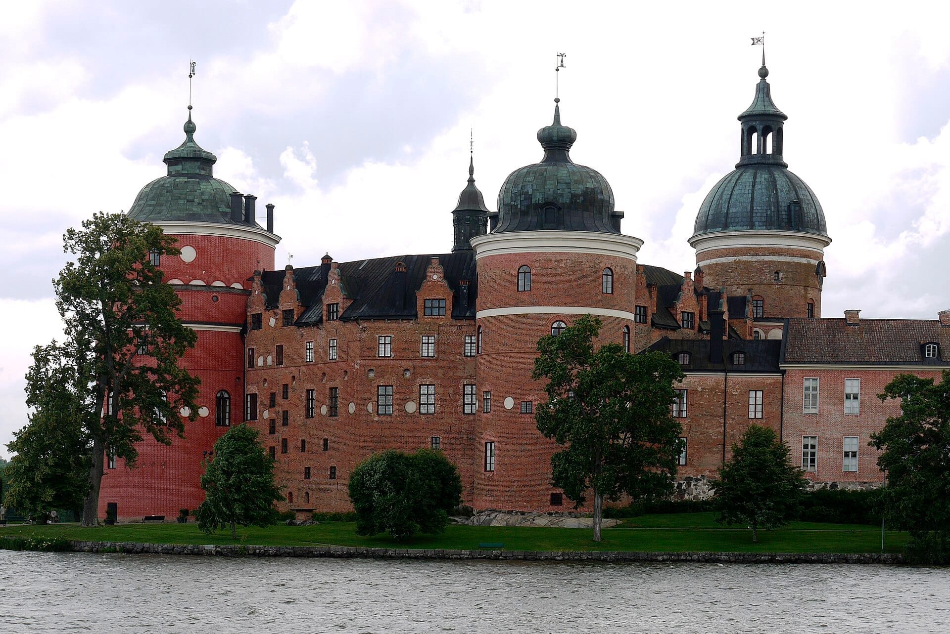 ปราสาท Gripsholm พร้อมหอคอยทรงกลมอันโดดเด่นและผนังอิฐสีแดง ล้อมรอบด้วยต้นไม้สีเขียวและหันหน้าสู่ทะเลสาบ Mälaren ใต้ท้องฟ้าที่มีเมฆปกคลุม