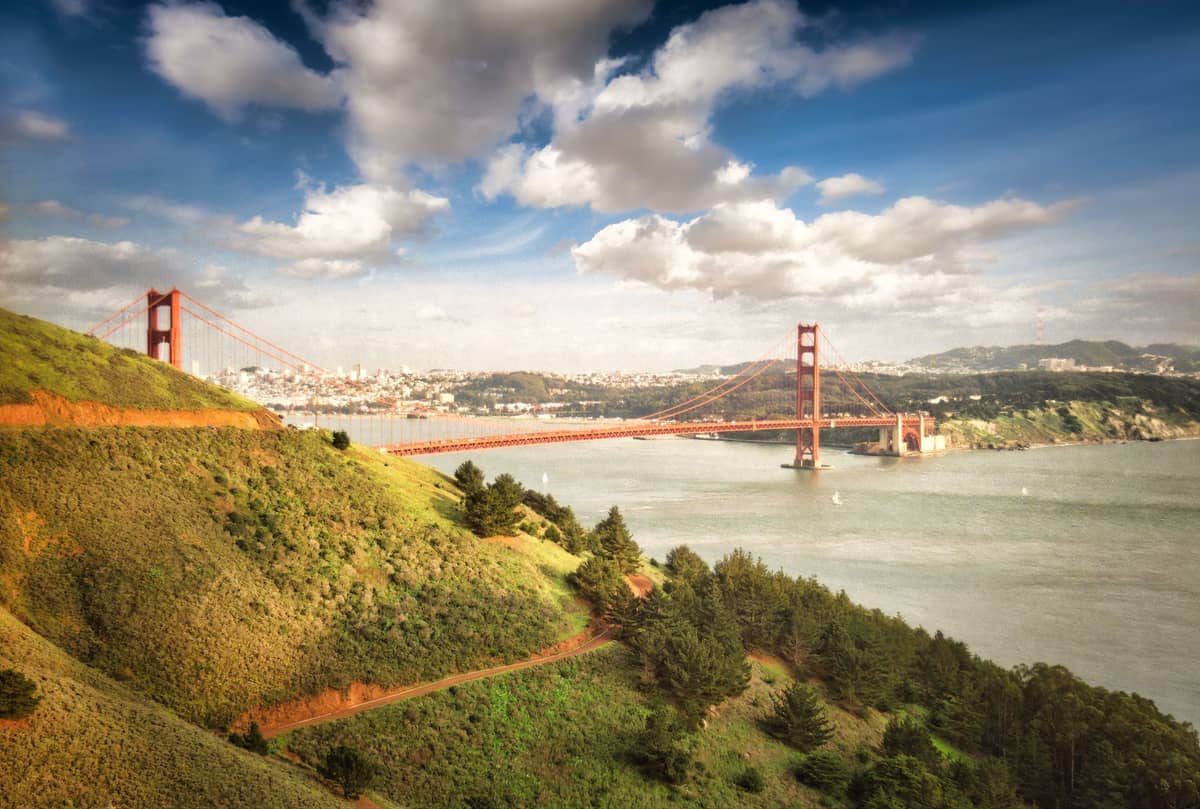 Vista panorámica desde los Marin Headlands con colinas verdes en primer plano, el Golden Gate Bridge a la derecha y San Francisco visible al otro lado de la bahía bajo un cielo parcialmente nublado.