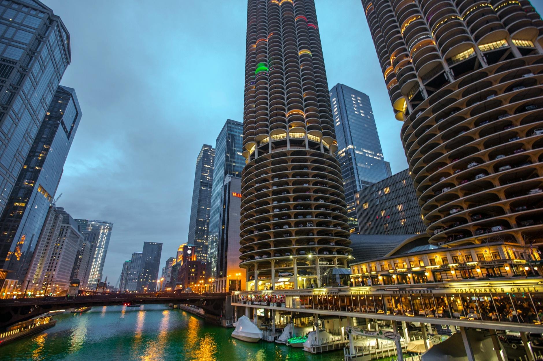 Vista serale delle due torri cilindriche di Marina City sul Chicago River, circondate da grattacieli, con le luci della città riflesse nell'acqua e l'animazione della riverwalk.