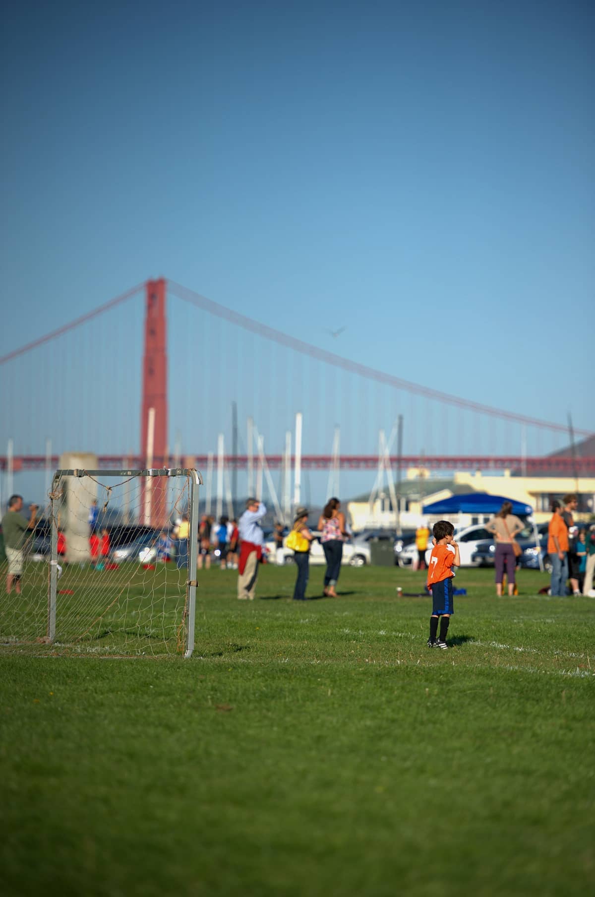 Niños jugando al fútbol en el campo de hierba de Marina Green con el Golden Gate Bridge y veleros visibles al fondo.
