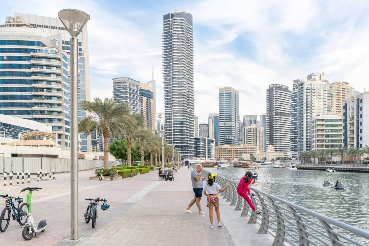 Die Promenade des Dubai Marina Walk mit Spaziergängern, modernen Hochhäusern, Palmen, dem Kanal und festgemachten Booten bei klarem Wetter.